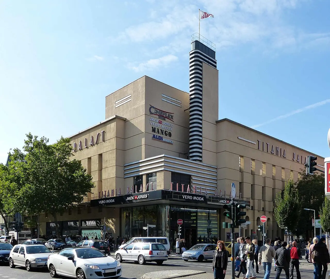 A large cream-coloured corner building with a striped tower and shop signs above a busy street.