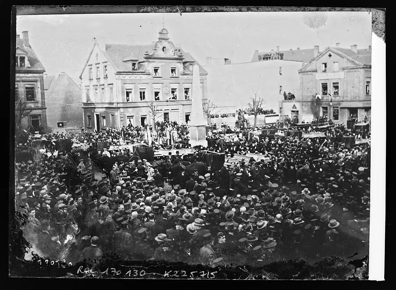 Seen from above, a dense crowd in hats and dark coats surrounds draped coffins on a town square lined with houses.
