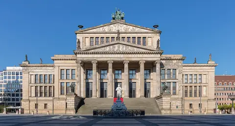 A classical building with a columned portico above a broad flight of steps, a statue in front, on a paved square