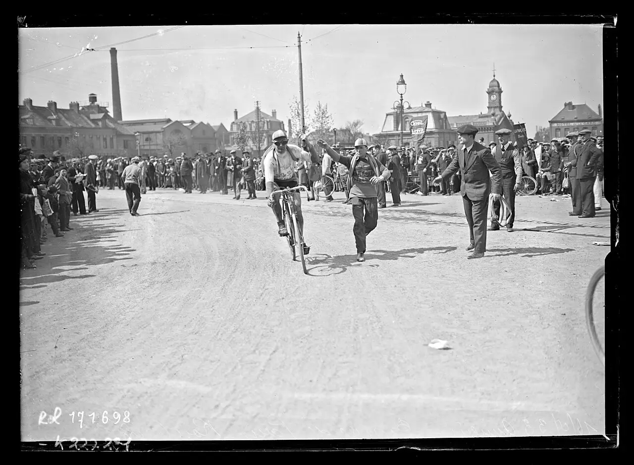 A racing cyclist rides across a wide town square while a man runs alongside and spectators line the edges.