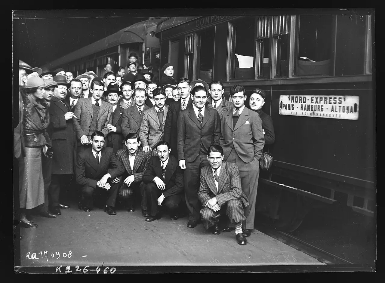 A group of young men in suits and ties, some crouching in front, posed with officials and onlookers on a station platform beside a sleeping car marked 'Nord-Express Paris–Hamburg–Altona'.