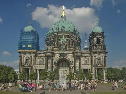 A large domed church with a green copper cupola and corner towers, with a fountain and people on the square in front