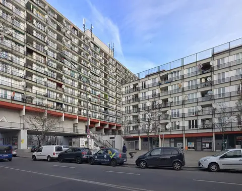 Two tall blocks of flats with rows of balconies and satellite dishes above a street lined with parked cars