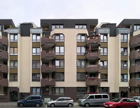 A six-storey apartment block with rows of projecting balconies and cars parked along the kerb