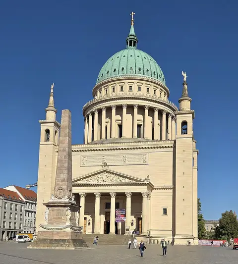 A domed neoclassical church with a green copper dome and a columned portico, with an obelisk in the square before it.