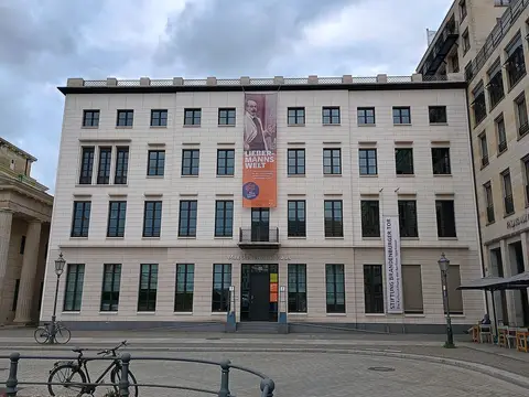 A pale five-storey building with tall windows on a square, a long exhibition banner on its front and the edge of a classical gate on the left