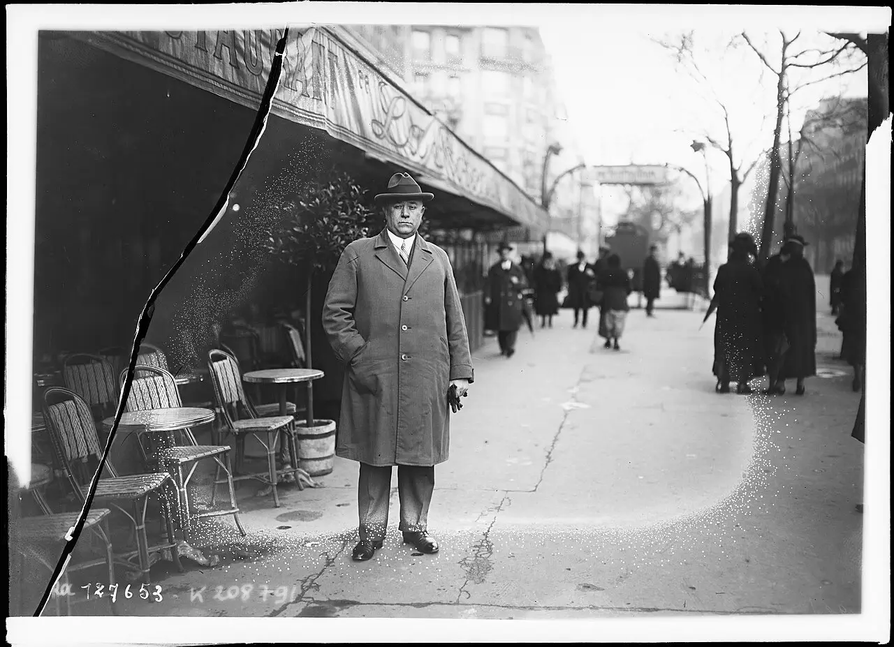 A man in a hat and long overcoat stands on a Paris pavement beside café tables, with pedestrians in the background.
