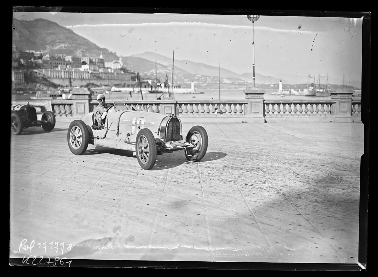 A racing car with the number 10 and its driver on a waterfront road, with the harbour and hills of Monaco behind.