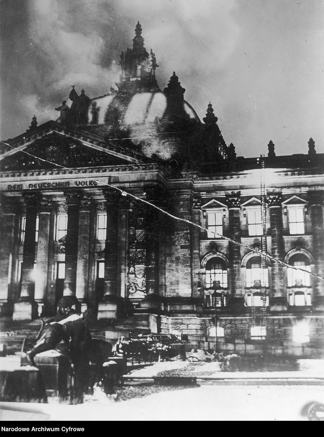 Night photograph of the Reichstag's columned west front, windows lit from inside, with dark smoke pouring from the dome.