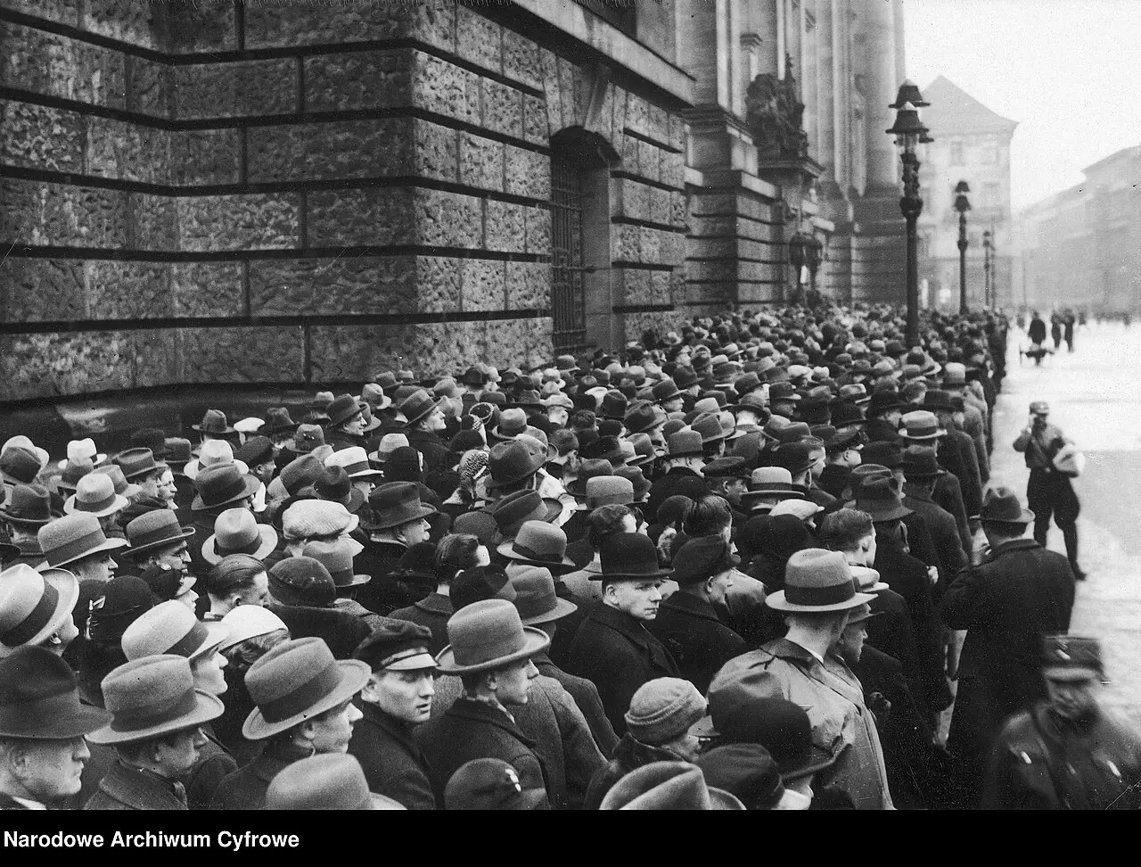 A dense crowd of men in hats packed along the rusticated stone wall of a large building, with lamp posts and a snowy street beyond.