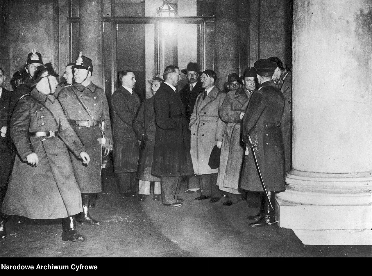 Night photograph of men in coats and hats and helmeted police officers in greatcoats gathered at a lit doorway beside a massive stone column.