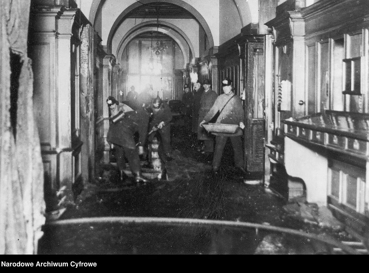 A vaulted, panelled corridor in which helmeted firemen carry a bucket and work with a hose that runs across the wet floor.