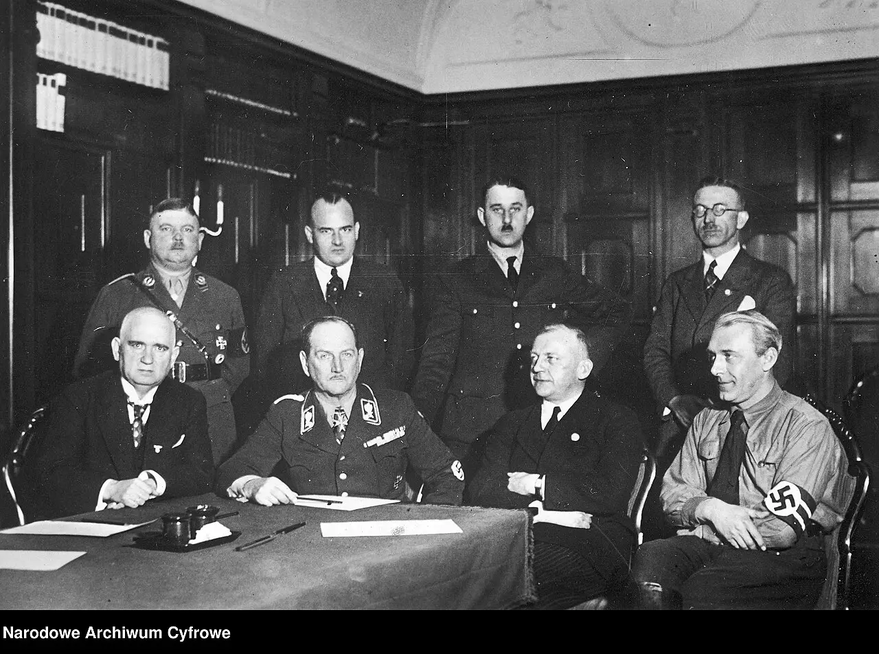 Eight men in a panelled room, four seated at a table and four standing behind them, several in uniform