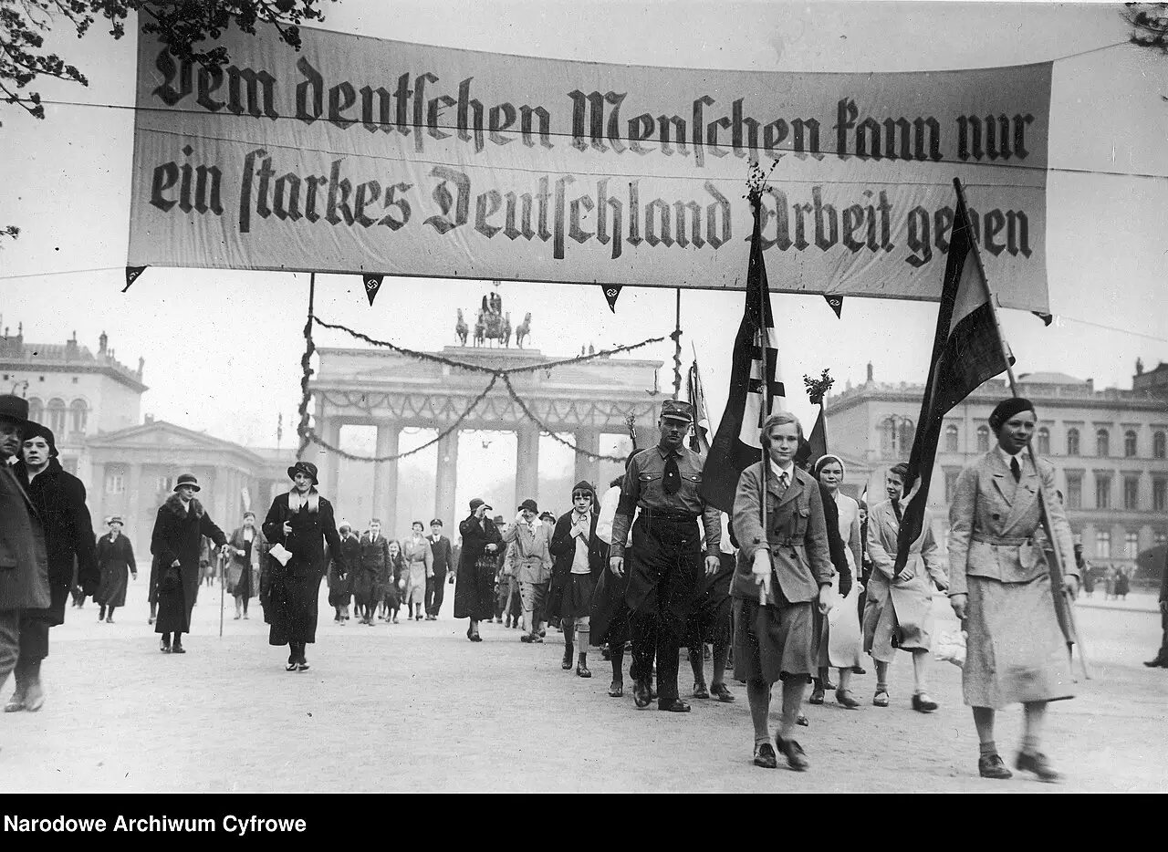 Young women carrying flags lead a march across a square under a large slogan banner, with the Brandenburg Gate in the background