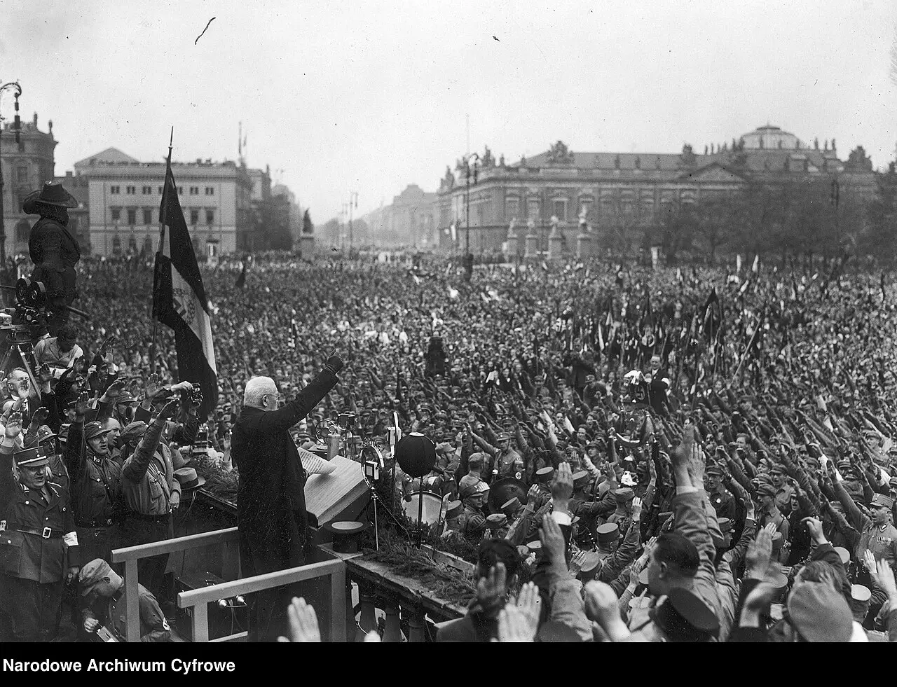 An elderly man in a dark coat raises his arm on a draped platform above a vast crowd giving the Nazi salute, with large public buildings behind.