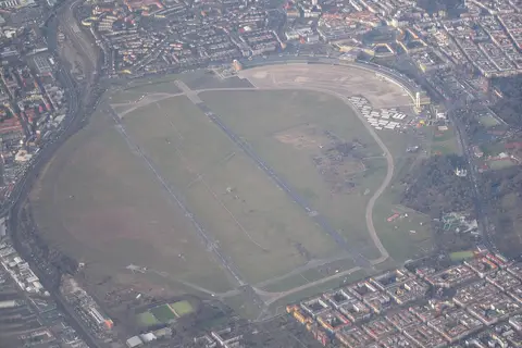 Aerial view of a vast oval open space crossed by two runways and surrounded by dense city blocks
