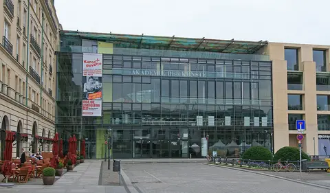 A glass-fronted building with the words Akademie der Künste across its facade, between two stone-clad buildings on a paved square