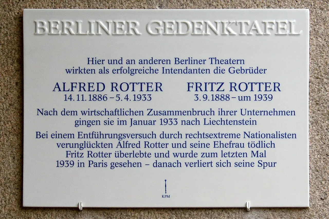 A white enamel memorial plaque with black lettering on a stone wall.