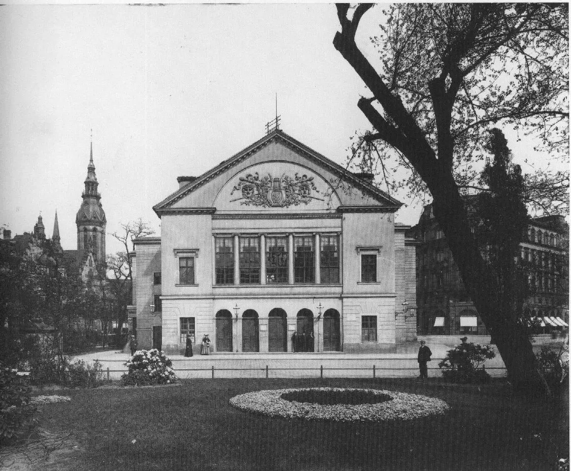 A classical theatre with a pedimented front and arched doors, seen across a garden with a flower bed