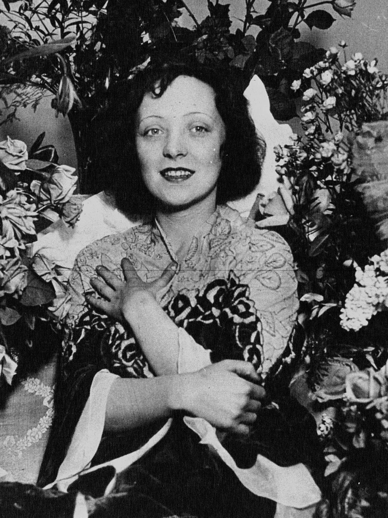 A smiling young woman with dark bobbed hair, surrounded by bouquets of flowers.
