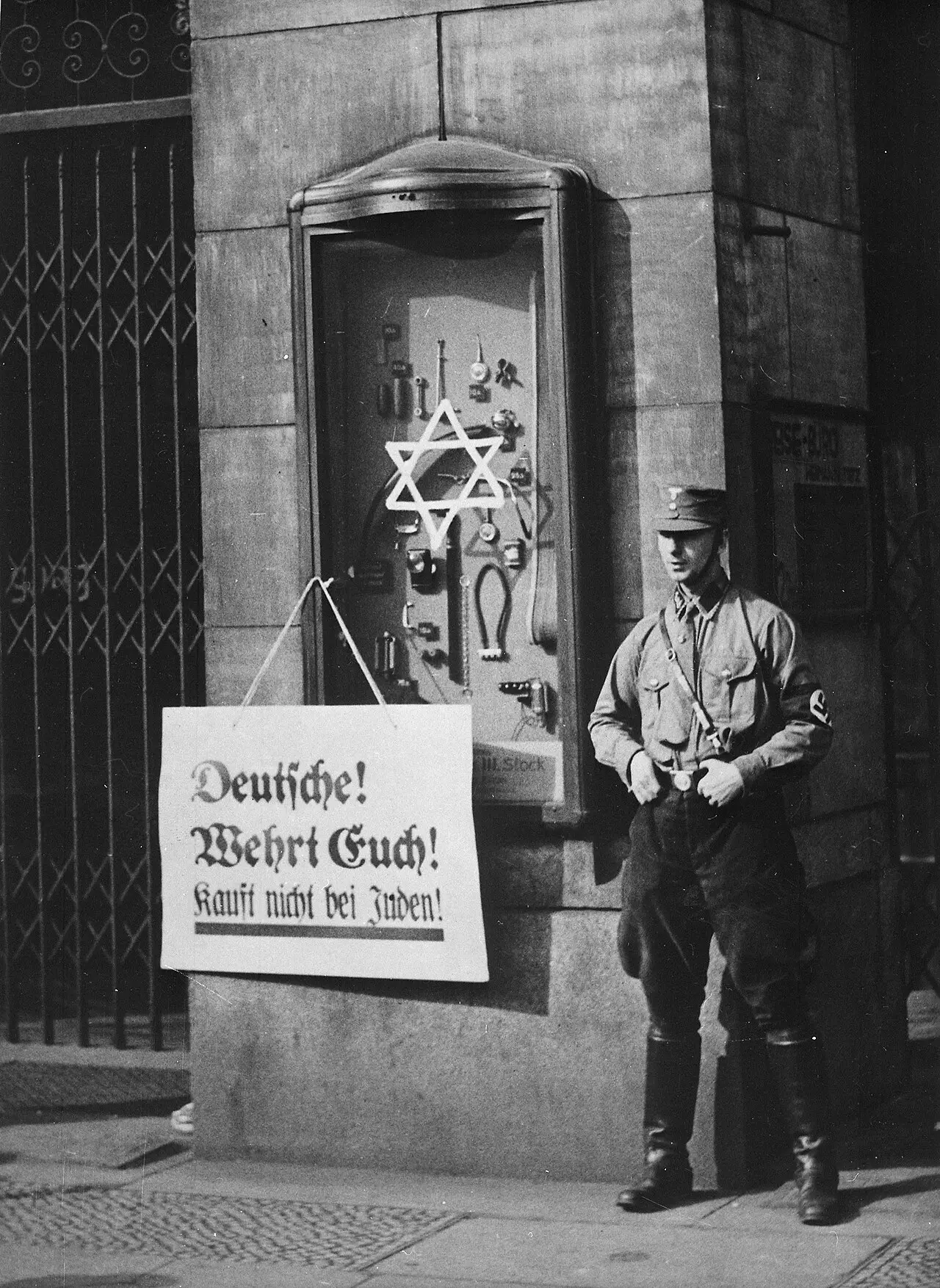 A uniformed SA man stands beside a shop display case on which a six-pointed star is painted; a placard with the boycott slogan hangs from the case.