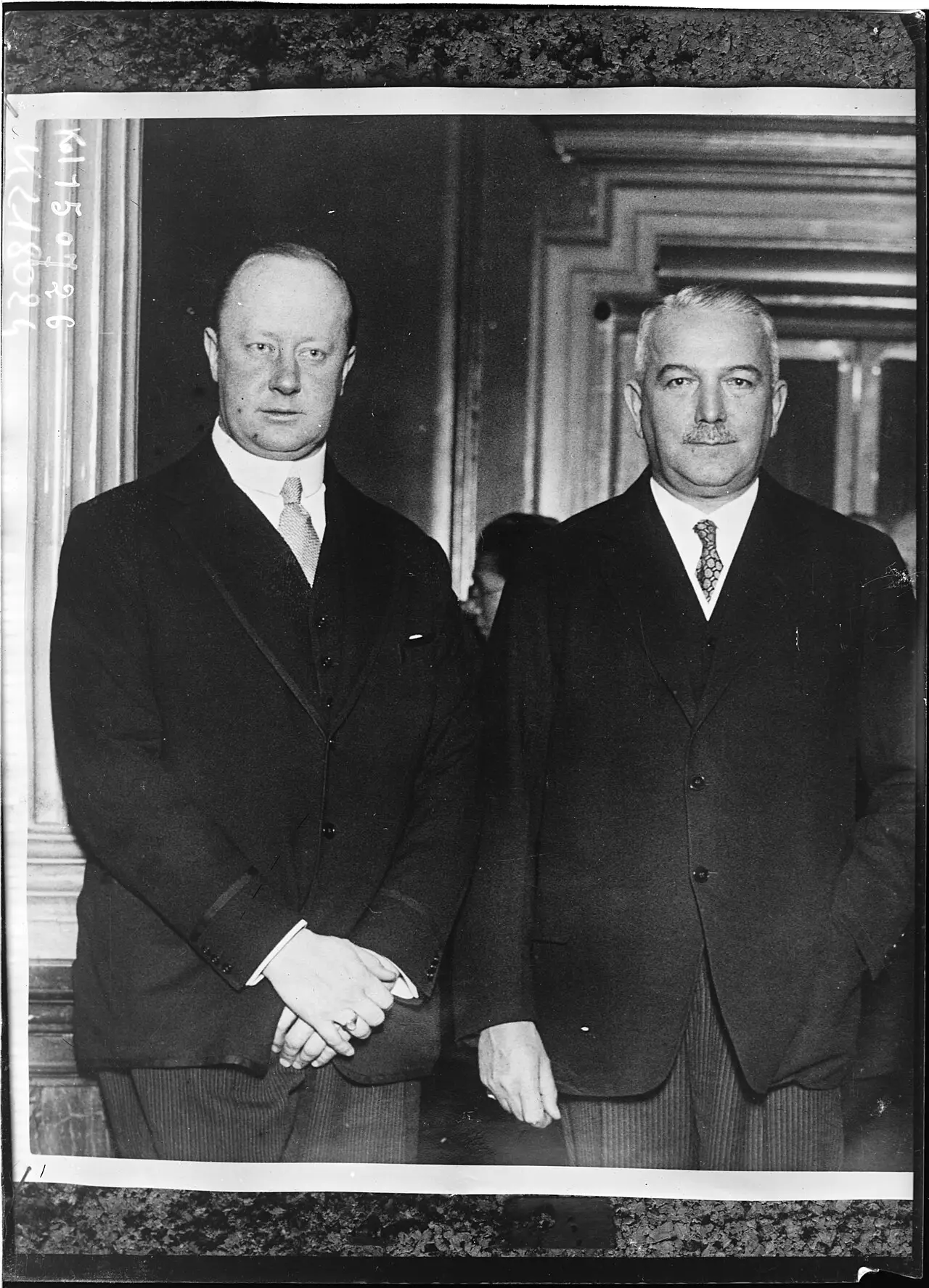 Two men in dark suits stand side by side in a panelled room: on the left a tall, heavily built younger man with his hands clasped, on the right an older man with grey hair and a moustache.