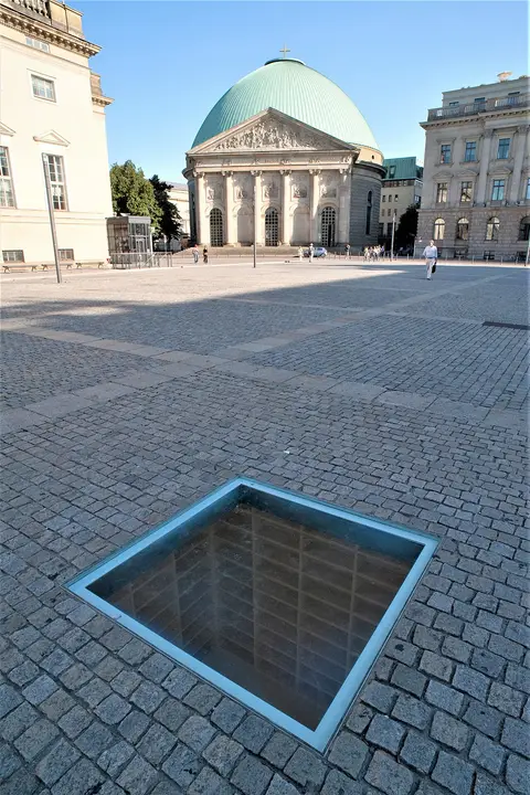 A square glass plate set into cobblestones shows empty bookshelves below; a domed church stands across the square