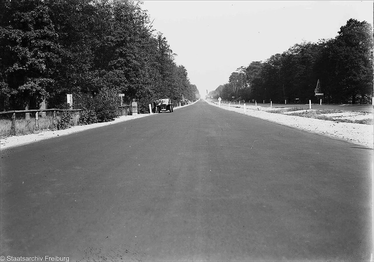 A broad, empty asphalt road running straight into the distance between woods, with a single car at the edge