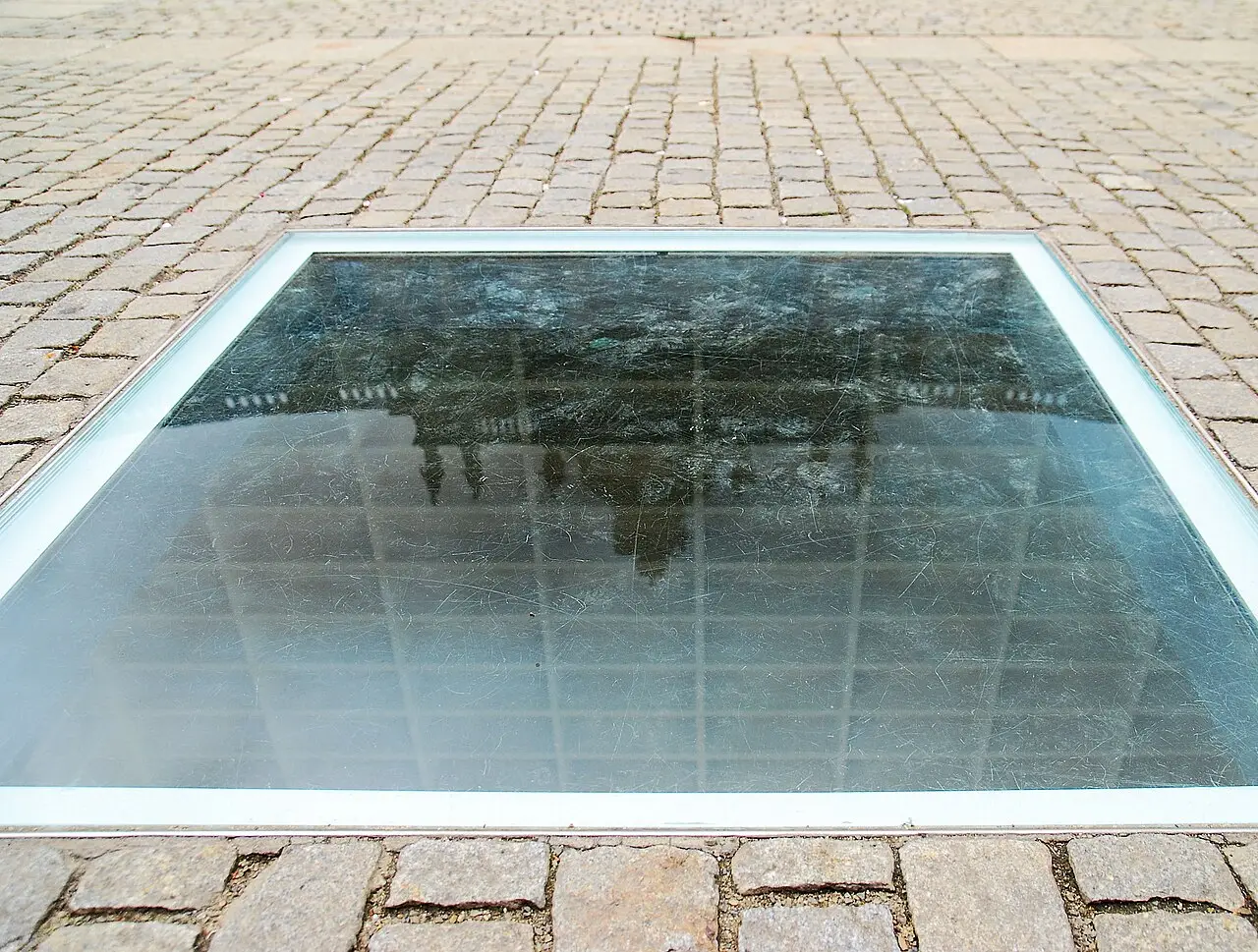 A square glass pane set into cobblestones, reflecting a building above faint white shelving below