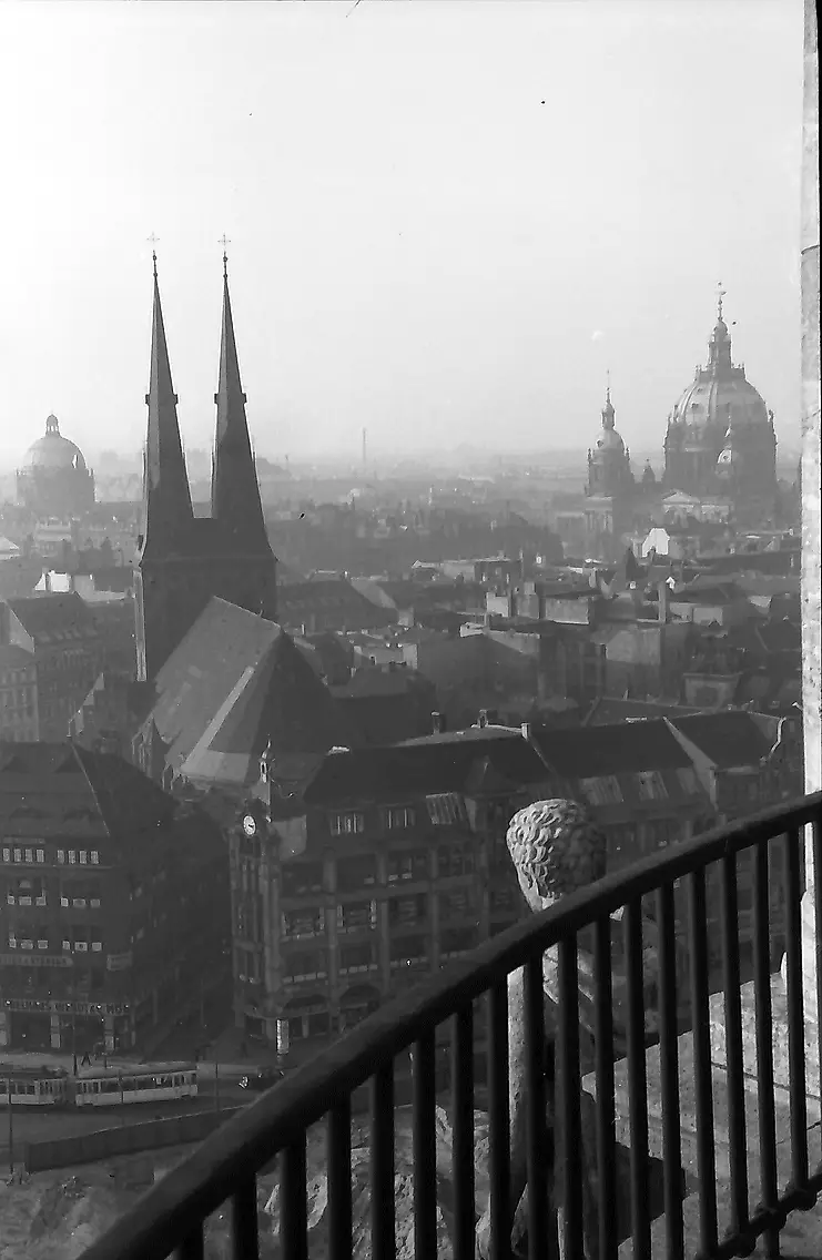 A view over Berlin rooftops from a high balcony, with a church with two tall pointed spires in the foreground and domes in the hazy distance.