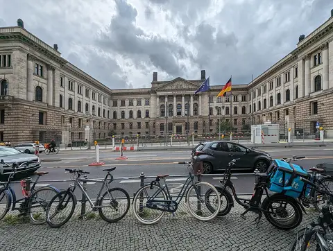 A long neoclassical stone building around a forecourt, with a pedimented centre and German and European flags, seen across a street with parked bicycles in the foreground.
