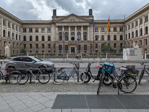 A long neo-classical building with a columned central pavilion behind a forecourt, seen across a street with parked bicycles