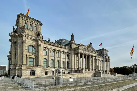 The sandstone west front of the Reichstag with its columned portico and corner towers, German flags and a glass dome behind, under a blue sky.