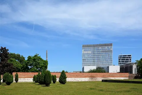 A long brick wall with a band of inscribed lettering along its base, behind a lawn with small conifers; modern office towers rise beyond.