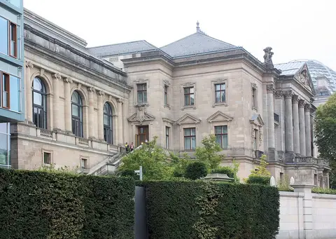 A two-storey sandstone palace with a mansard roof and a columned wing, behind a tall clipped hedge