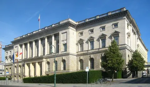 A large neo-Renaissance stone building with a columned centre, flags and a hedge in front