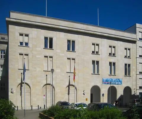A plain four-storey limestone building with arched ground-floor openings, flagpoles with German and European flags, and parked cars in front.
