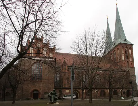 A large brick church with two tall green spires, seen across a small square with bare trees and a seated bronze figure.