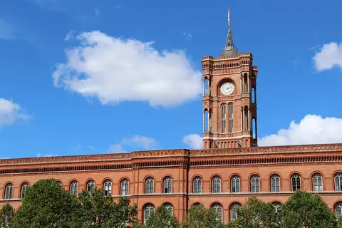 The upper storeys and clock tower of a long red-brick town hall with rows of arched windows, under a blue sky with white clouds, trees in front.
