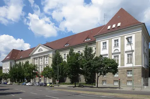 A long three-storey building with a steep red-tiled roof and a tall corner pavilion, behind trees on a quiet street