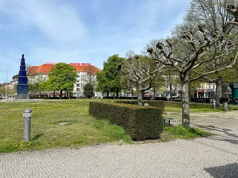 A broad green square with trees and clipped hedges, a tall blue obelisk at left and apartment buildings around the edge.