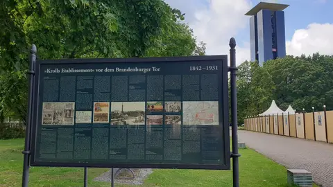 A large dark information board with text and pictures stands on a lawn beside a path; a tall bell tower rises behind the trees