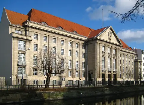 A long sandstone office building with a steep red-tiled roof and a pediment, behind railings on a canal bank
