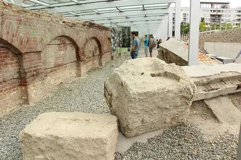 Old brick walls with arched recesses and fallen stone blocks on gravel beneath a glass canopy, with visitors reading display panels