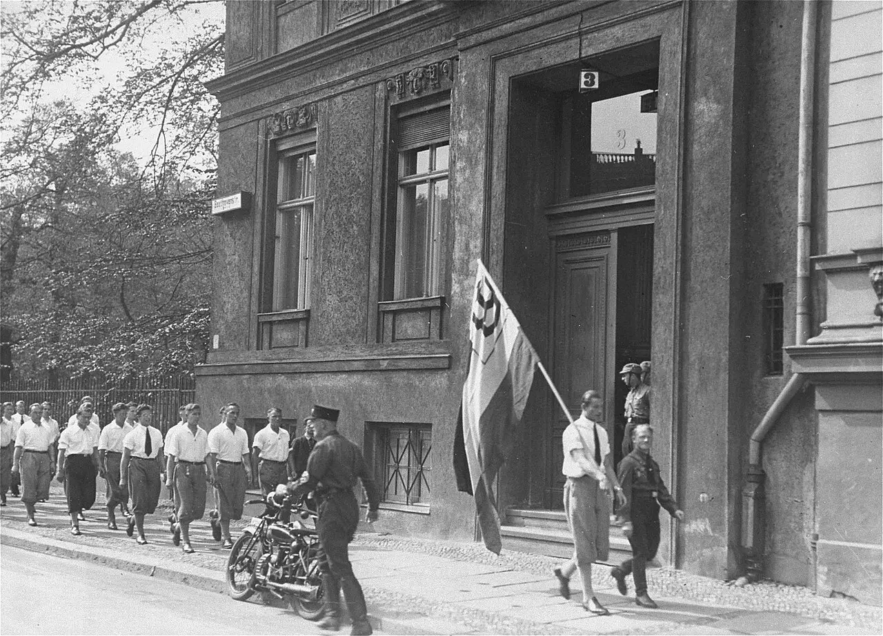 Young men in white shirts drawn up in ranks in front of a villa, with a lorry and a banner