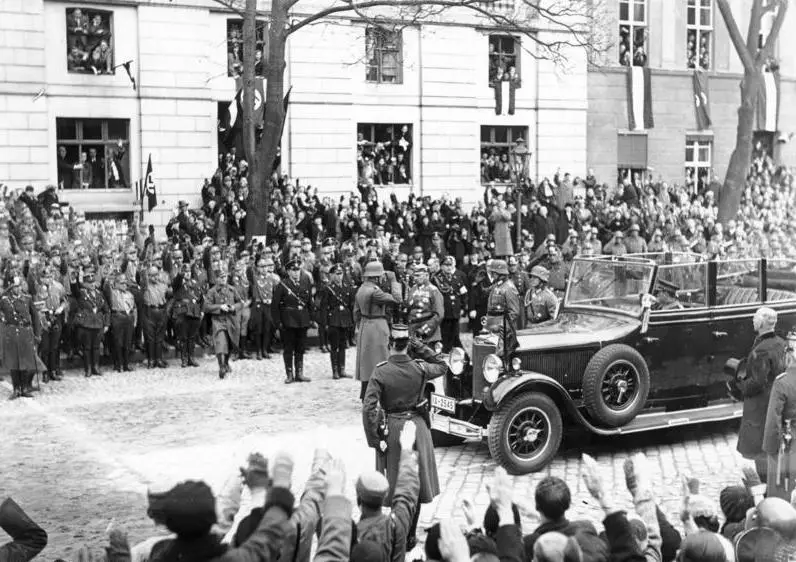 An open touring car on a cobbled street before ranks of uniformed men and a dense crowd, with spectators leaning from the windows of the buildings and raised arms in the foreground.