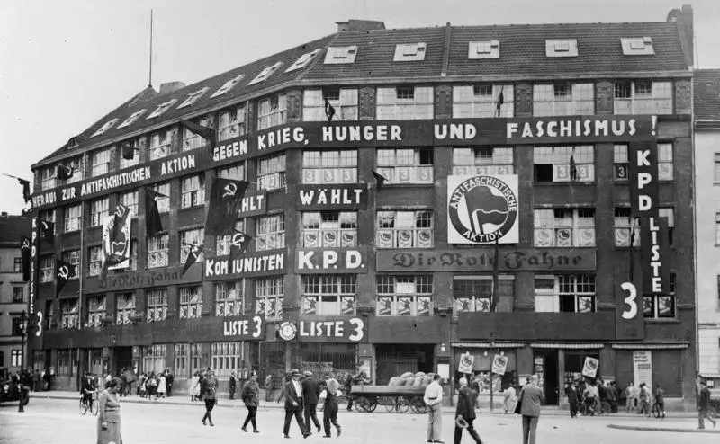 A five-storey corner building covered in banners and posters, with the Antifascist Action emblem, hammer-and-sickle flags and the words 'KPD Liste 3'; pedestrians and a handcart in the street below.