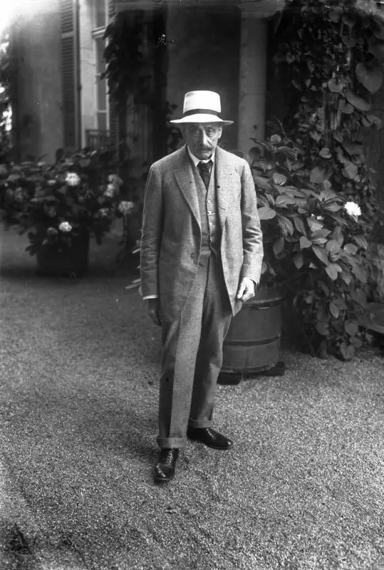 An elderly man with a white moustache in a light suit and hat standing outdoors