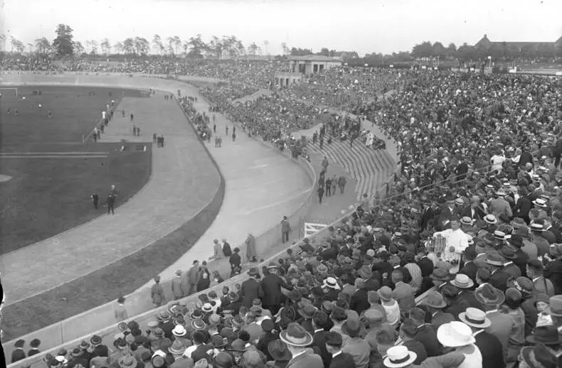 A packed grandstand of spectators in hats beside a running track and football pitch, with trees behind.