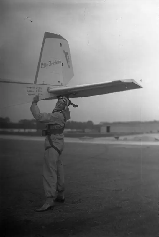 A woman in a flying suit and leather helmet stands on an airfield holding up the tailplane of a light aircraft; the fin is lettered 'Elly Beinhorn'.